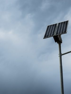 Solar panel on street lamp post with light on and cloudy sky.